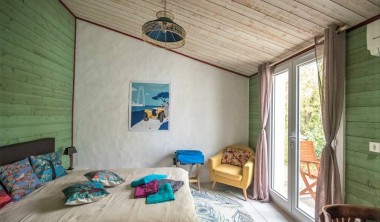 Chambre avec un mur blanc et deux murs en lambris vert, lit double et fauteuil, portes vitrées donnant sur un salon de jardin en bois. Chambre - Les Merlettes des Sables